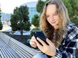 Teen redhead girl standing on urban street skyscrapers background wearing headphones using smartphone iPhone 13 listening to music, audio book, travel guide or podcast while walking in big city. photo