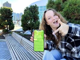 Teen redhead girl standing on urban street skyscrapers background wearing headphones using smartphone iPhone 13 listening to music, audio book, travel guide or podcast while walking in big city. photo