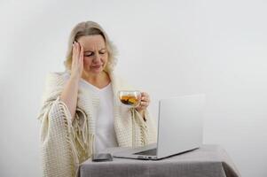 Sad unhappy exhausted sick woman suffering from common cold or flu fever wrapped in scarf, plaid and blanket, feeling unwell, laptop , touching forehead, holding mug and drinking warm tea photo