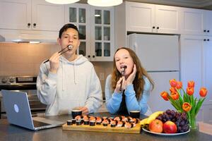 teenagers boy and girl eating sushi at home in the kitchen watching a movie on a laptop flowers grapes apples beautiful environment white room first date communication meeting friends photo