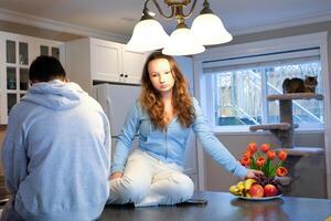 teenagers sit on table in kitchen girl offended upset pouted boy turned back on phone looks gadget eat fruit year in background near window adolescence quarrel of discontent offended at each other photo