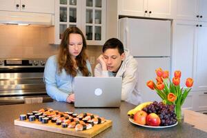 Mira a ordenador portátil apoyarse terminado enfocado pensar Mira adolescentes haciendo deberes en el cocina ordenado Sushi reunirse después colegio primero fecha reloj película gastar hora juntos chico niña 13 14 15 años antiguo foto