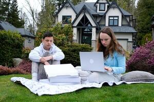 young teenage couple sits on white blanket girl holds laptop in hands she is busy boy is bored near three boxes of knitting needles Expectation and reality failed date you need to do your homework photo