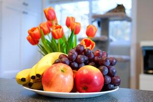 on a white plate apples grapes and bananas bright red tulips with a yellow rim close-up in the background a cat on a cat and a tree near the window kitchen breakfast photo