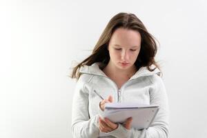 Portrait of attractive optimistic woman wearing sports suit standing with paper notebook and pen in hands, looking at camera with smile. Indoor studio shot isolated on gray background. photo