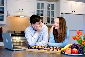 teenagers in kitchen looking after girl with loving eyes on table laptop sushi fruits flowers beautiful first date invite to visit friend smile look into eyes Boy and girl talking and eating sushi photo