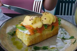 Typical but trendy breakfast close up shot, knife cut poached egg on top of egg Benedict meal, we see yellow liquid yolk inside. Stylish food with salmon and hollandaise sauce, served at trendy cafe photo