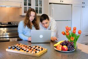 set up a laptop repair teenagers prepared to watch sushi on table tulips fruits in kitchen celebrate March 8 valentine's day birthday movie attentively focused look at gadget cooking holiday fun photo