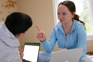 teenage girl explaining something to friend boy waving forefinger pointing at laptop with white screen work study fun learning homework tutor help study together at home after class come visit meeting photo