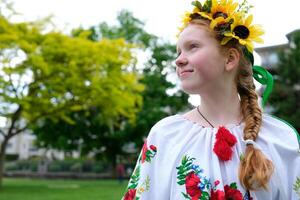 young red-haired girl in a wreath of sunflowers outdoors in park garden in embroidered Ukrainian vyshyvanka shirt with red braid with ribbons in hair Ukraine Peaceful Time of happiness joy and peace photo