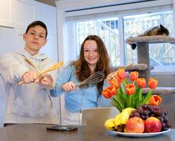 kitchen utensils in the hands of a teenage boy and girl in kitchen dancing celebrating having fun together in background cat sleeping by the window on a cat tree fruit on the table nice day date fun photo