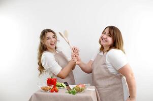 two women two friends fight on wooden spoons smile laugh in the frame play have fun on the table knife vegetables nuts aprons of the same color on a white background cooking cooking grocery store photo