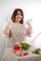 alegre hembra cocinero es sentado a el mesa con manojo de verdura, demostración preparado comida y Okay signo.femenino cocinero demostración preparado comida y Okay firmar foto