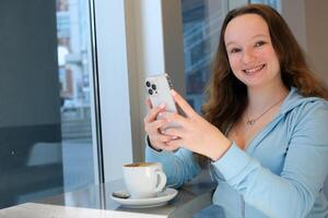 Beautiful cute young teen girl in the cafe, using mobile phone and drinking coffee smiling the girl looks into the frame laughs in her hands the phone sit by the window In a bright cafe office photo