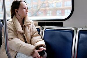 There is space for text Ukrainian fair haired European girl sits in skytrain and she listens to music in front of her holds the phone She is in a white blouse and blue ripped jeans she is in a tunnel photo