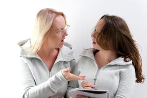 mom and daughter doing homework, woman wearing glasses, girl trying to write, they are laughing, smiling, spending pleasant time, explaining to child, surprise, pointing with hand at notepad, notebook photo