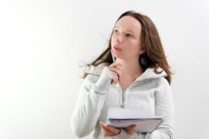 young teenage girl studying at school, lost in thought, biting her pen in wind, leafing through sheets of notebook, sitting by the window close-up in classroom on white background, one young woman photo