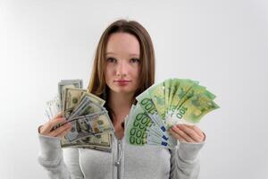 rich teenager girl waving a fan of 100 euros and 100 dollar bills standing on a white background smiling in a sports gray sweater loose brown hair surprise calm rich child photo