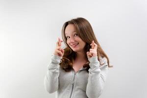 girl is experiencing wonderful expectations, crossing two fingers near face of both hands, waiting for everything to come true, smiling gently, straightening hair on white background in studio photo