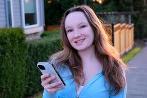 Portrait of happy hipster woman typing by mobile phone outdoors. Closeup cheerful girl walking with smartphone in urban background. Smiling lady holding cellphone in hands outside. photo