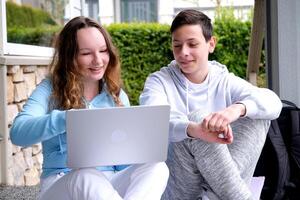 sit outside on porch whisper in ear have fun laugh doing homework boy and girl heterosexual friends teenager using laptop and tablet for online schooling from home together, quarantine concept. photo
