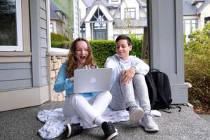 sit outside on porch whisper in ear have fun laugh doing homework boy and girl heterosexual friends teenager using laptop and tablet for online schooling from home together, quarantine concept. photo