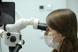 Brunette woman dentist in a white coat and a medical protective mask works with a modern microscope in the office. Operations in the dental clinic. photo