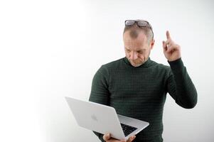 a man in a green sweater, an adult, raised his finger up, put glasses on his forehead, looks in surprise at a laptop on a white background Student using laptop isolated on white photo