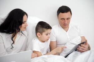 banner family husband wife and child sitting watching movie on laptop on bed relaxing together white clothes chatting online with relatives mom dad and son laughing smiling rejoicing happy life photo