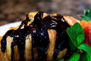 a lot of dark chocolate topping poured on a fresh croissant with mint and strawberries on a white plate cook at home who likes chocolate chocolate too much is never too sweet too much food photo