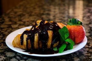 a lot of dark chocolate topping poured on a fresh croissant with mint and strawberries on a white plate cook at home who likes chocolate chocolate too much is never too sweet too much food photo