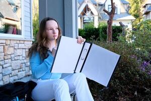 Portrait of creative pensive little girl teenager school child caucasian smart pupil stands alone in room at home on window background holding folder with paper comes up with idea plan writing note photo