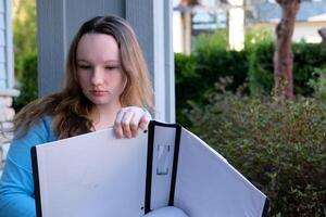 teenage girl with a big white folder in hands crosses out something with pencil writes draws creative person artist young teens outdoors sit by porch have fun holding pen wrong learning doing homework photo