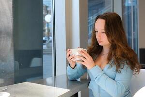 joven niña adolescente o Bebiendo café Dom es brillante ella mira a taza grande vaso ventanas restaurante suelto pelo Espere para amigo oficina sueño trabajo descanso estudiar Universidad cantina cafetería foto