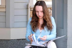 young girl teenager sits on porch in hands with a folder, a magazine and pencil, do homework, solve problems, wait at the door, a beautiful woman, young white jeans, a briefcase, think, work, study photo