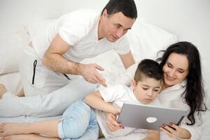 people, family and technology concept mother, father and little boy with tablet pc computer in bed at home or hotel room. Happy family spend time together lying under white blanket on bed at bedroom photo
