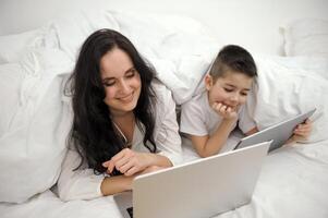 chico, y su madre es trabajando en un ordenador portátil computadora, el pequeño chico poner el tableta en su mano y convertido a su ocupado madre atentamente, falto su madre a pagar atención a a él. foto