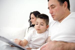 family, fatherhood, technology and people concept happy father and little son with tablet pc computer sitting in bed at home mom woman working in the background online on computer laptop photo