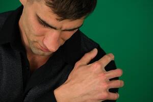 a guy left shoulder hurts on a green background chromakey in black clothes man holds his hand close-up suffering from pain sprain dislocation muscle tension needs massage with copy space for text photo