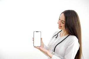 Woman doctor or nurse wearing a lab coat and stethoscope pointing to a blank mobile phone with a pleased smile close up over a grey background photo