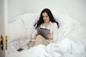 beautiful black-haired woman sits on white sofa near a duvet in hands with tablet corner of room works plays communicates nearby mobile phone social networks Internet Call friend pleasant emotions photo