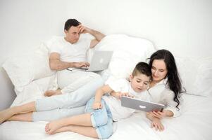 mamá entretiene hijo en tableta blanco ropa uno cama descanso apartamentos idilio gastar hora juntos ser solo con tú mismo familia moderno comunicación en línea Internet social redes computadora comunicación foto