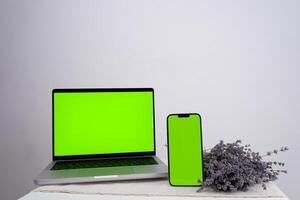 blank mobile phone while using laptop with blank green screen on table Mockup image of Laptop mobile blank green screen in vertical position isolated on green background, Concept device mockup. photo