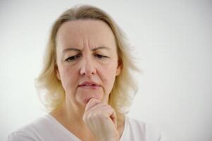 Photo of thoughtful woman bites lower lip, looks with dreamy expression aside, wearing stylish beige hoodie, stands against white wall. Copy space for your text