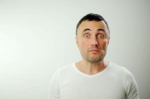 hombre es sorprendido él tirado fuera su ojos elevado su Cejas fuertemente y arrugado su frente él es en un blanco camiseta en un blanco antecedentes en su cara sin afeitar oscuro pelo grande azul ojos foto