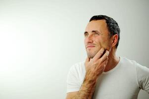 toothache going to the doctor Adult man in white t-shirt white Background Holding on to his cheek It hurts Veins popped out of man's arms The unshaven man has black hair and gray hair at the temples photo