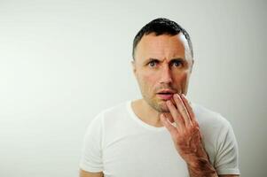 toothache going to the doctor Adult man in white t-shirt white Background Holding on to his cheek It hurts Veins popped out of man's arms The unshaven man has black hair and gray hair at the temples photo