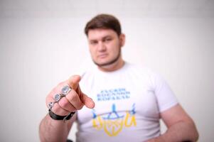 Handsome adult broad shouldered man shows a frame with his finger He is blurry. On the fingers there is a clear ring with coat of arms of Ukraine. He has a white T-shirt. It says Russian ship go on photo