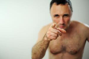 man with a naked torso and hairy arms With one hand held at the temple on the head Adult unshaven handsome man shows his finger Open wide blue eyes white background studio Advertising space for text photo