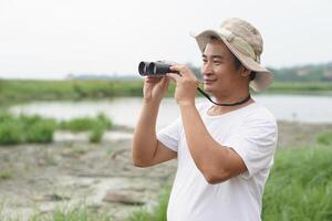 Handsome Asian man ecologist is surveying nature at the lake, holds binoculars.Concept, nature exploration. Ecology study. Pastime activity, lifestyle. Man explore environment photo